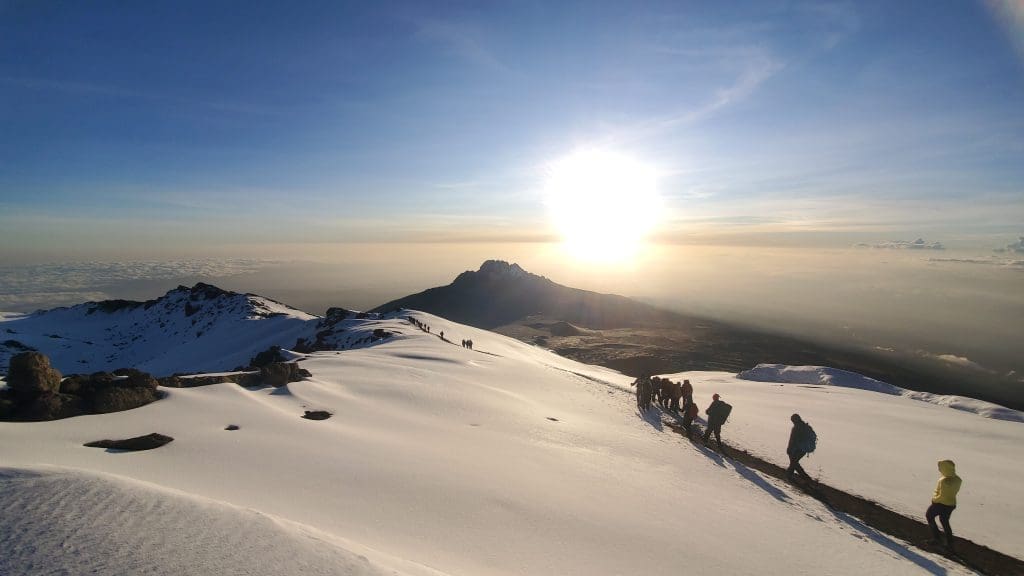 Climbers trek across the snow-covered slopes near the summit of Mount Kilimanjaro at sunrise, with the sun shining brightly over Mawenzi Peak.