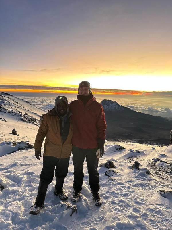 Mountain guide and tourist standing together on a snowy slope near the summit of Mount Kilimanjaro at sunrise, with Mawenzi peak and clouds in the background.