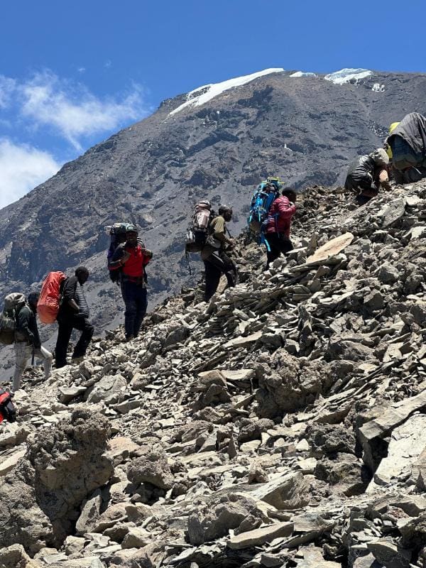 Group of climbers ascending a steep rocky slope on Mount Kilimanjaro with towering peaks and patches of ice in the background.